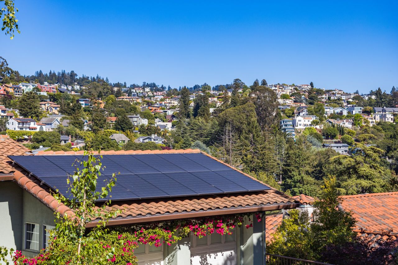 who-we-are Solar panels on a suburban home, surrounded by lush greenery and a sunny blue sky.